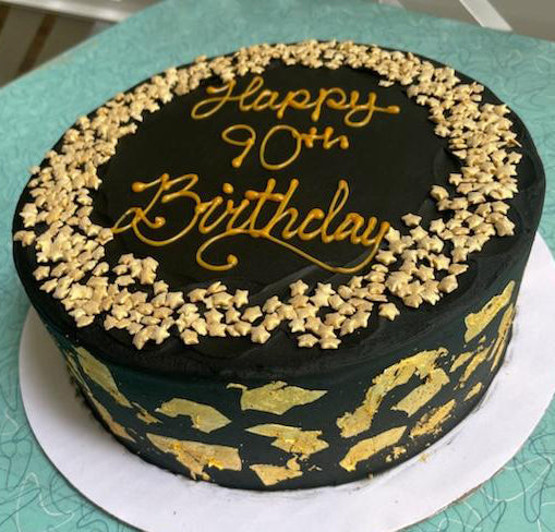 Black birthday cake with gold decorations and 'Happy 90th Birthday' text on a green surface.