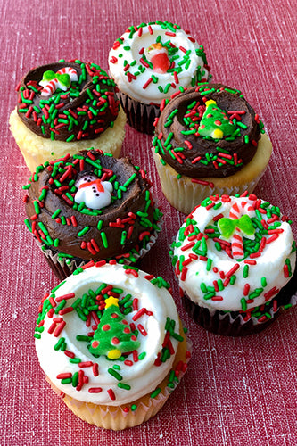 Six assorted Christmas-themed cupcakes, some with chocolate and some with vanilla frosting, decorated with red and green sprinkles, and edible holiday candies like Christmas trees, snowmen, and candy canes.