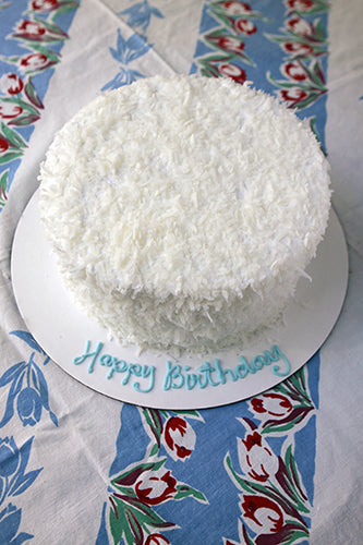 White coconut cake with Happy Birthday written in blue icing on the cake board, on a floral tablecloth.