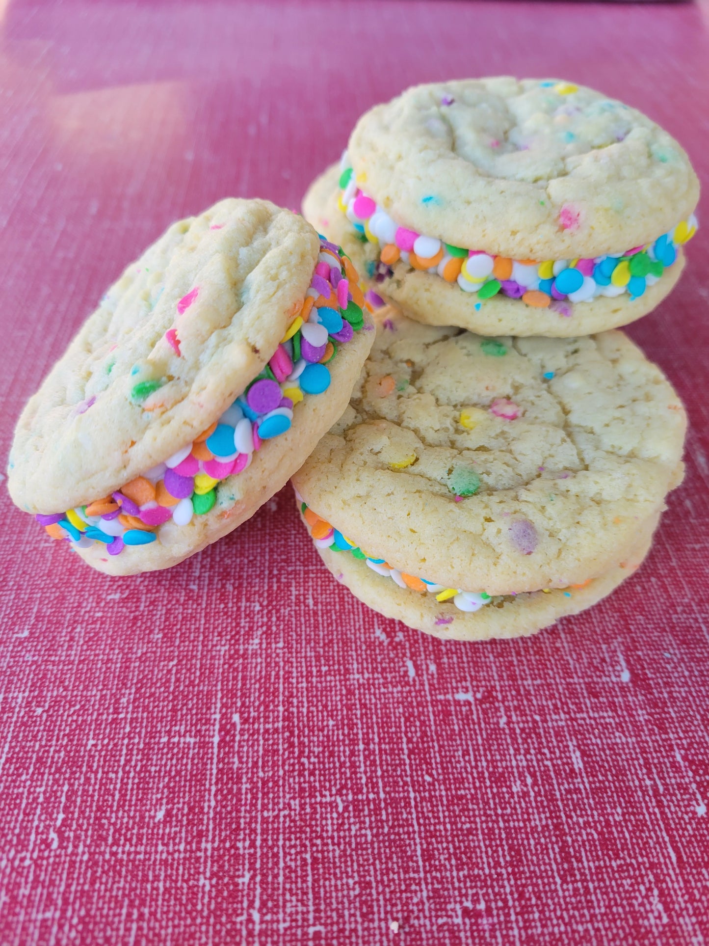 Numerous confetti cookie sandwiches on a red table, each with a creamy white filling, and rolled in colorful confetti garnish. 