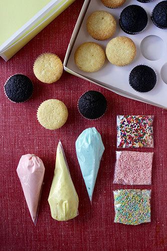Overhead view of a cupcake decorating kit with plain and chocolate cupcakes, piping bags with colorful frosting, and bags of sprinkles on a red surface.