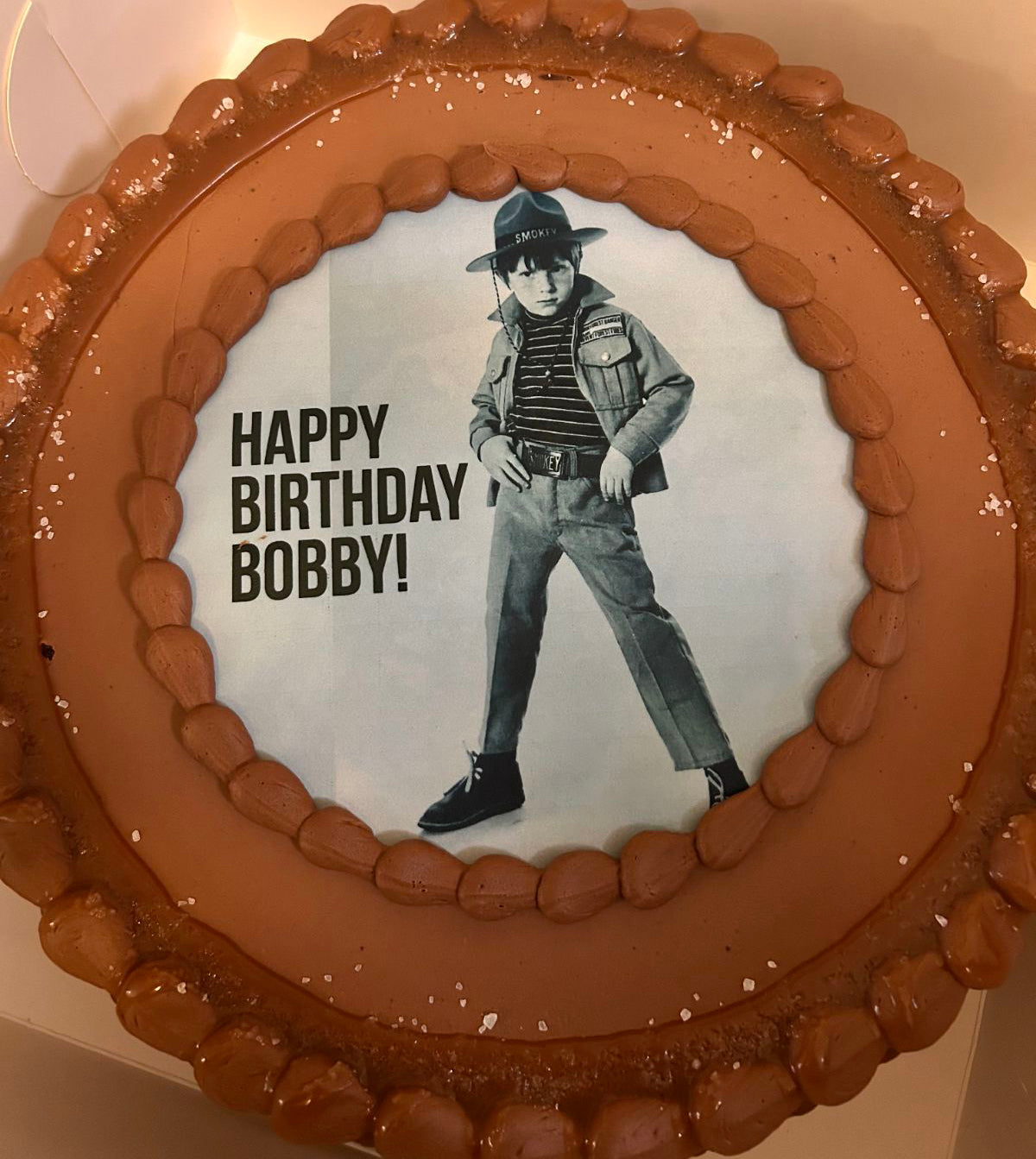 A round cake with brown icing featuring a printed image of a character and the text 'HAPPY BIRTHDAY BOBBY!'