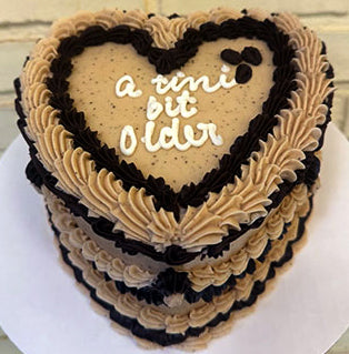 A heart-shaped chocolate cake with espresso buttercream and chocolate accents, decorated with the text 'a tiny bit older' on top, and garnished with espresso beans.