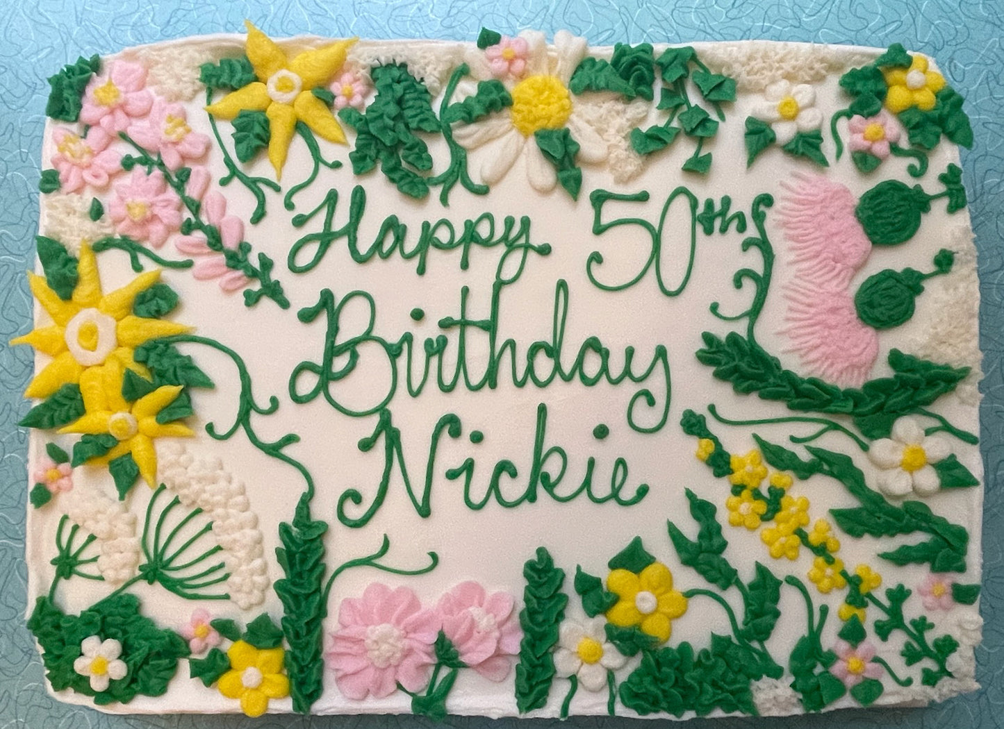 Birthday cake with floral decorations and 'Happy 50th Birthday Nickie' text on a blue background.