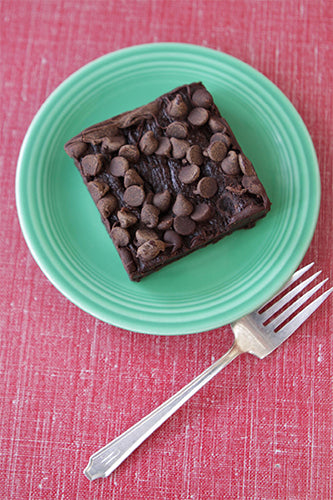 A gluten-free chocolate fudge brownie on a green plate with a fork, featuring chocolate chunks on top.