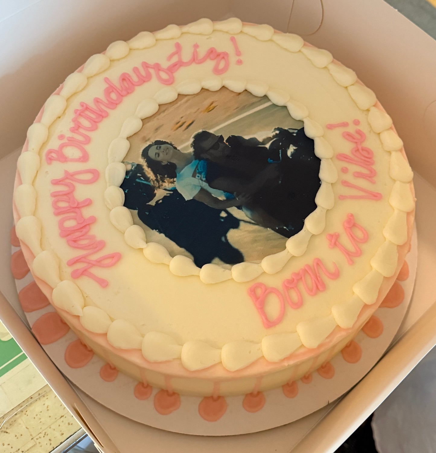 Round cake with white frosting, pink text, and a photo of two people in the center.