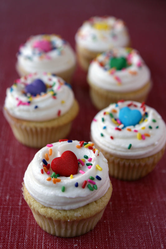 Decorative cupcakes with white frosting, colorful sprinkles, and heart decorations on a red surface.