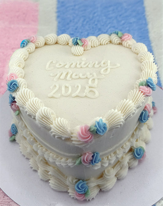 A heart-shaped cake with white buttercream frosting, decorated with pastel pink and blue rosettes and green leaves.