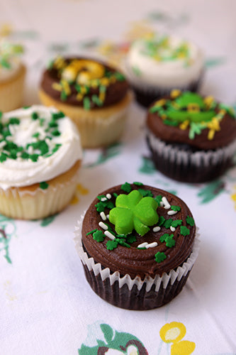 A collection of St. Patrick's Day themed cupcakes, including chocolate cupcakes with green frosting and shamrock decorations, and vanilla cupcakes with white frosting and green sprinkles, arranged on a festive tablecloth.