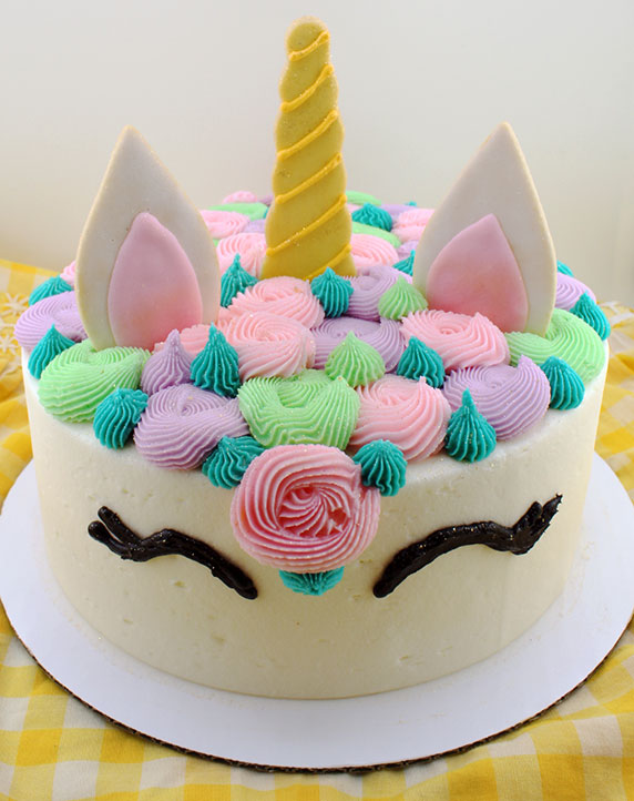 A decorated cake designed to look like a unicorn with white 'fur' rosettes, pink ears, and a yellow horn, presented on a yellow checkered background.