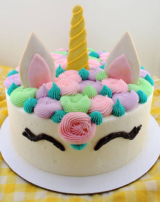 A decorated cake designed to look like a unicorn with white 'fur' rosettes, pink ears, and a yellow horn, presented on a yellow checkered background.