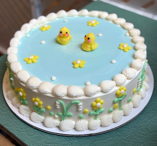 A decorated cake with white buttercream frosting and blue buttercream, adorned with yellow daisy-like flowers and small sugar ducks.