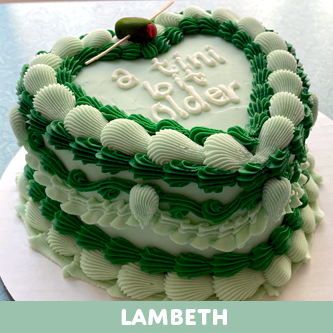 A heart-shaped cake decorated with light and dark green Lambeth-style frosting, topped with an olive on a pick and white icing text saying "a bit tiny older."