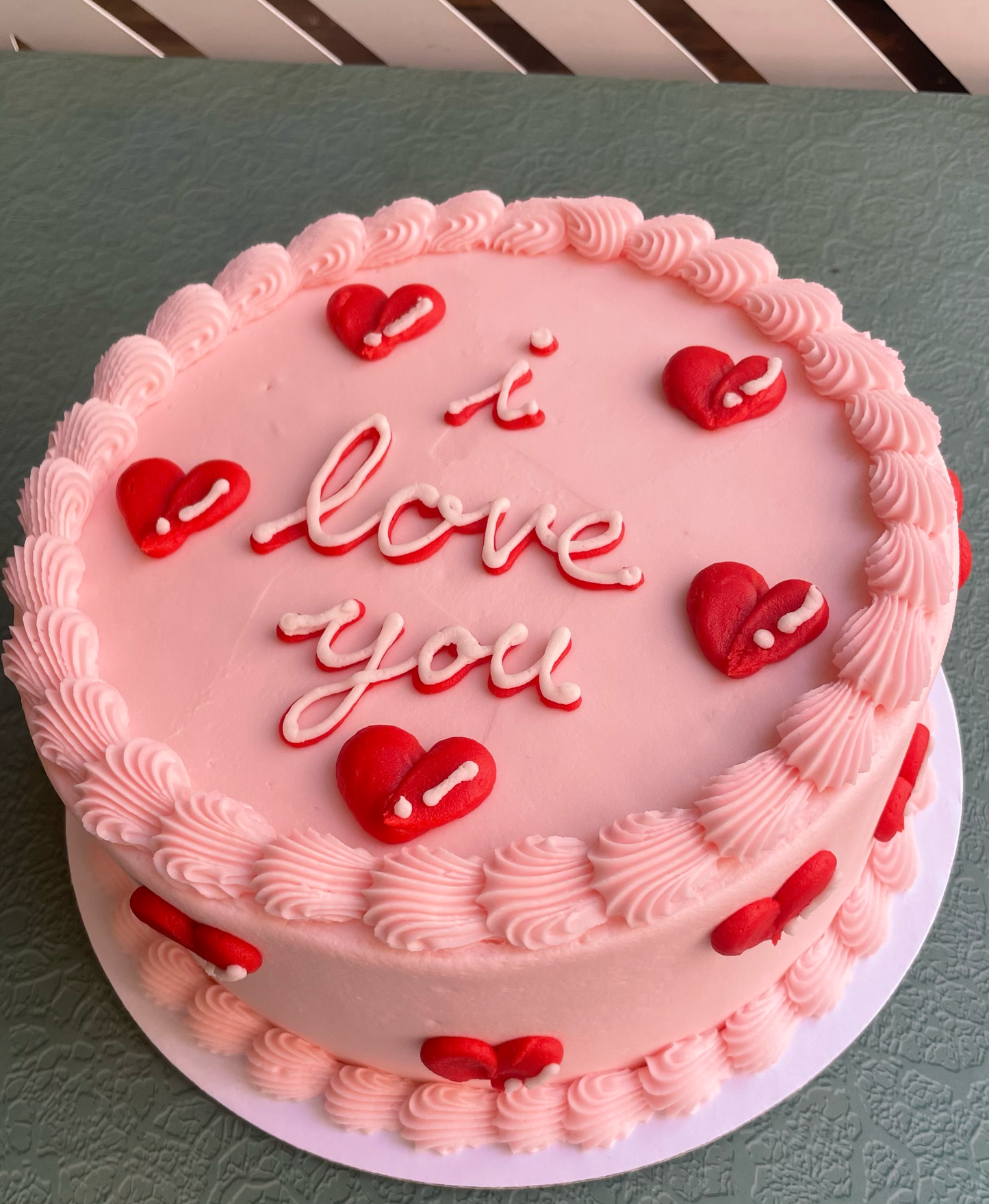 A frosted cake with pastel pink buttercream and decorated with puffy red bubble hearts and the words 'i love you' in cursive on top.